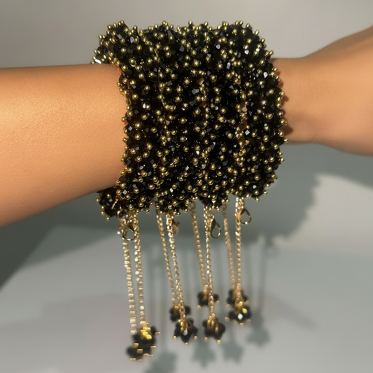A close-up of a person's wrist wearing a stack of wide black beaded bracelets with dangling gold-tone chains and bead clusters.