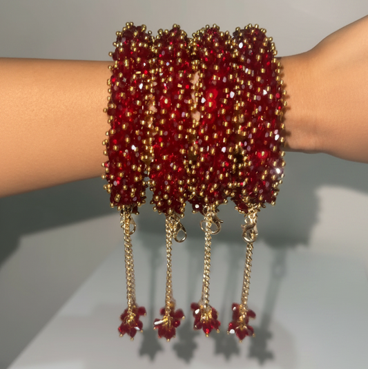 A close-up of a person's wrist wearing four wide red beaded bangles with gold-toned chain tassels and dangling floral charms, set against a plain light gray background.